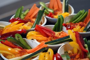 A close-up view of multiple small bowls filled with hummus and a colorful arrangement of sliced vegetables. The bowls contain vibrant strips of yellow, red, and green bell peppers, along with orange carrot sticks and green cucumber slices, all artfully arranged and sprinkled with a dusting of paprika. 
