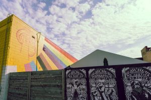 A rainbow mural is painted on the side of a yellow brick building. The wall stands against a partly cloudy sky, with a white tent and wooden fence in the foreground. Black-and-white protest art posters with messages: “Minneapolis United” and "We Rise Up Together" and "No Justice No Peace" 
