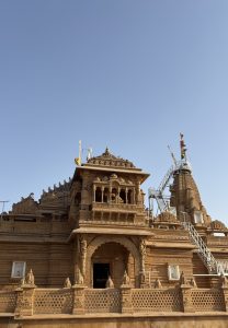 A beautifully carved sandstone Hindu temple with intricate architecture, domes, and balconies, featuring flags atop its spires and a metal staircase leading to the shikhara, under a clear blue sky.