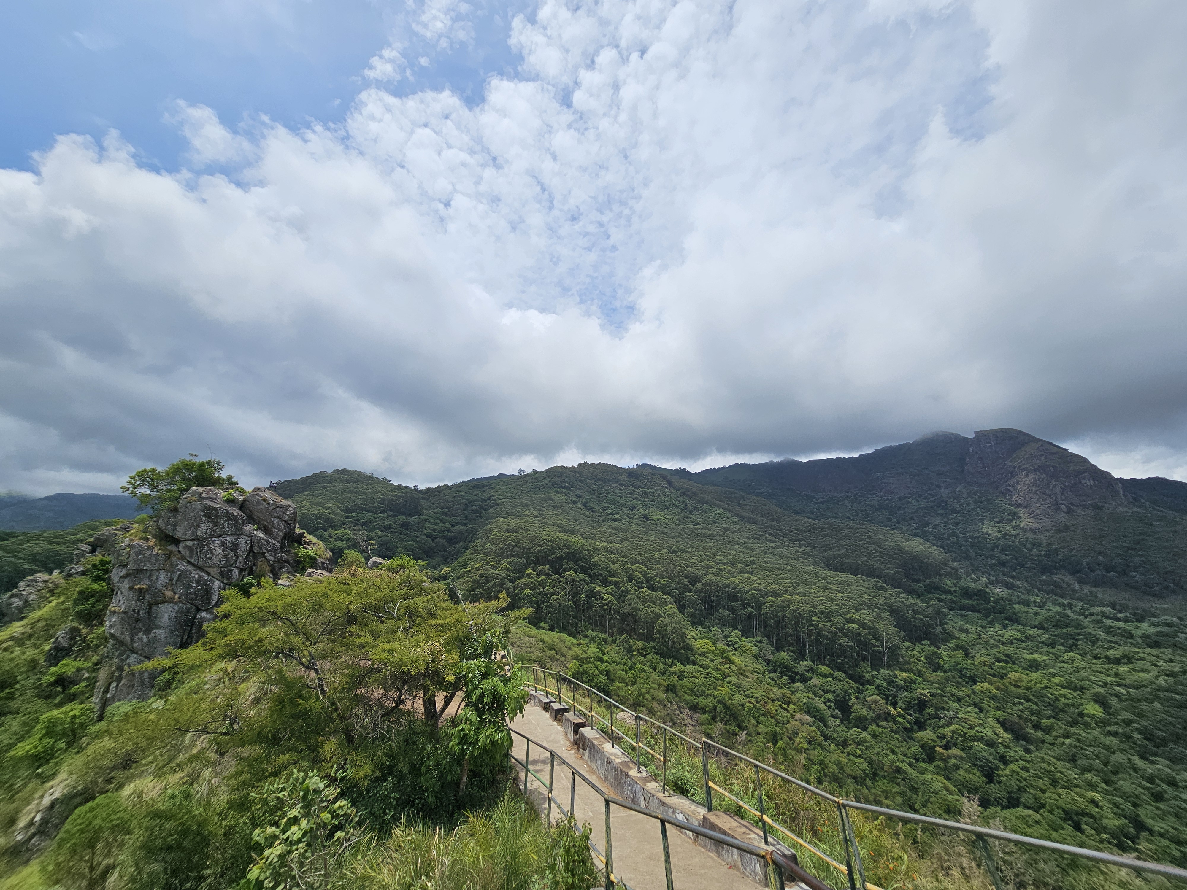 A scenic view from Needle Rock View Point in the Nilgiris, Tamil Nadu, India. It features a pathway with railings leading to a panoramic view of lush green mountains and a cloudy sky.