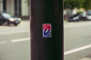 An Uno reverse card in the colors of a bisexual pride flag on a black street pole. In the background there are blurry cars and trees.