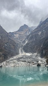 A panoramic view of a rugged mountainous landscape featuring steep, rocky cliffs and a glacier. Below, a tranquil turquoise lake reflects the surrounding terrain.