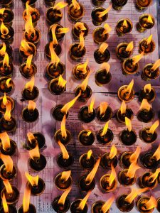 A top-down view of numerous small oil lamps with flickering flames, arranged in a grid pattern on a surface that appears to have printed sheets underneath.
