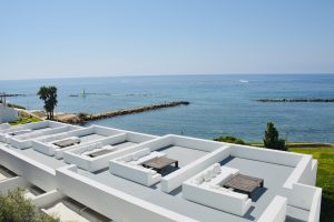 A modern rooftop terrace with white lounge seating and wooden tables overlooking the blue sea. In the distance, breakwaters, a sailboat, and palm trees along the coast are visible. Taken in Paphos, Cyprus.