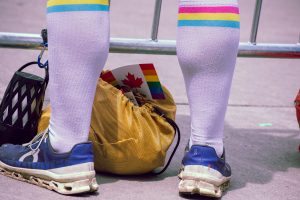 A person wearing white knee-high socks with rainbow stripes and blue running shoes stands beside a yellow drawstring bag. A Canadian flag with rainbow borders peeks out from the bag, resting on the pavement near a barricade during a Pride event. 