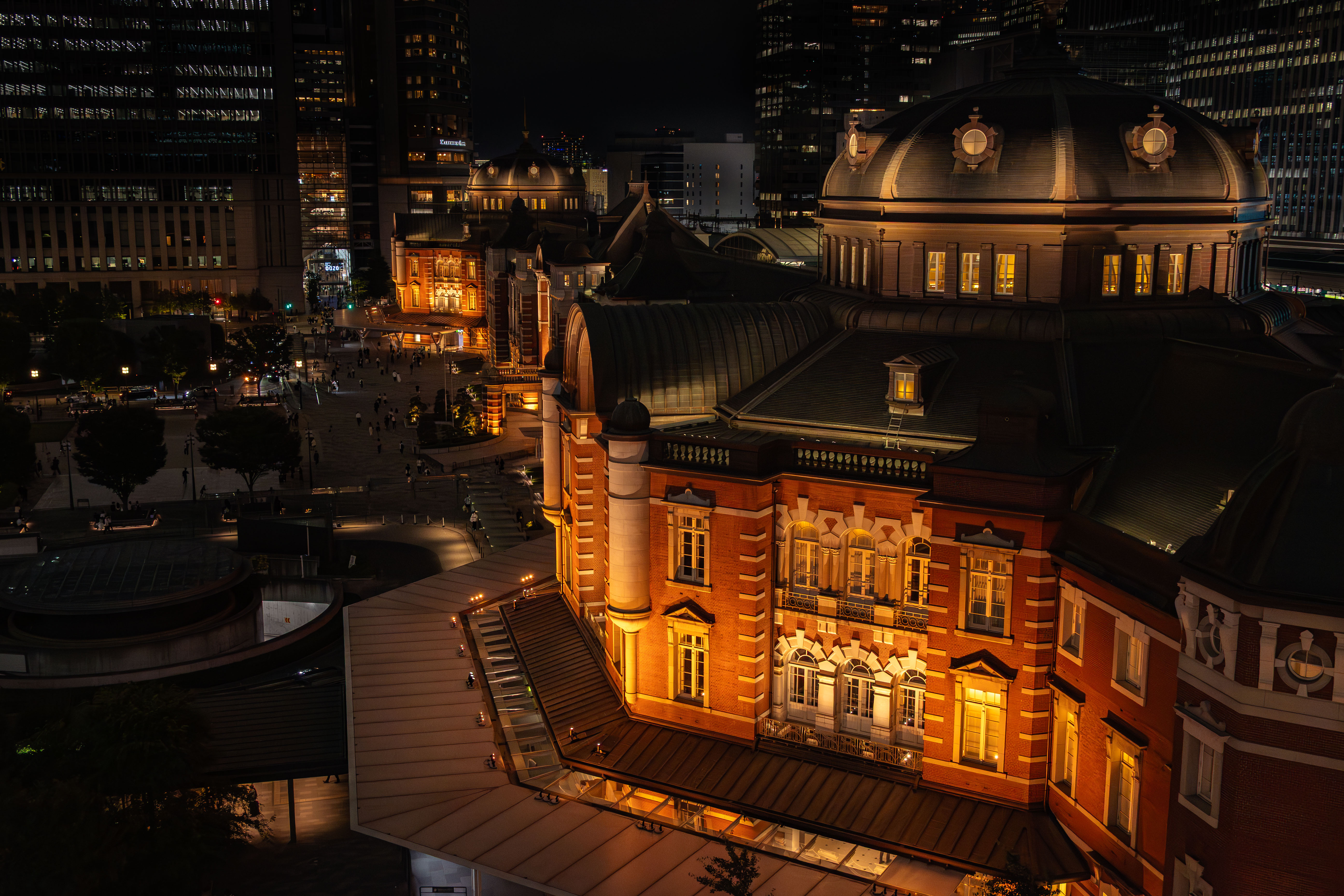 Marunouchi exit of JR Tokyo Station at night. View from the rooftop of a nearby facility. The bricks are lit up in orange, and the Yaesu skyscrapers can be seen in the background.