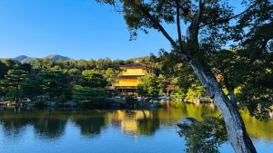 A serene landscape featuring a golden pavilion situated on the edge of a tranquil pond, surrounded by lush green trees. 