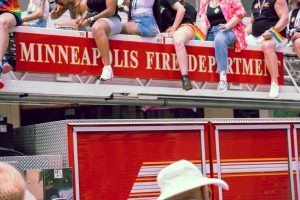 People sit on top of a Minneapolis Fire Department ladder truck during a Pride parade, smiling and waving rainbow flags.
