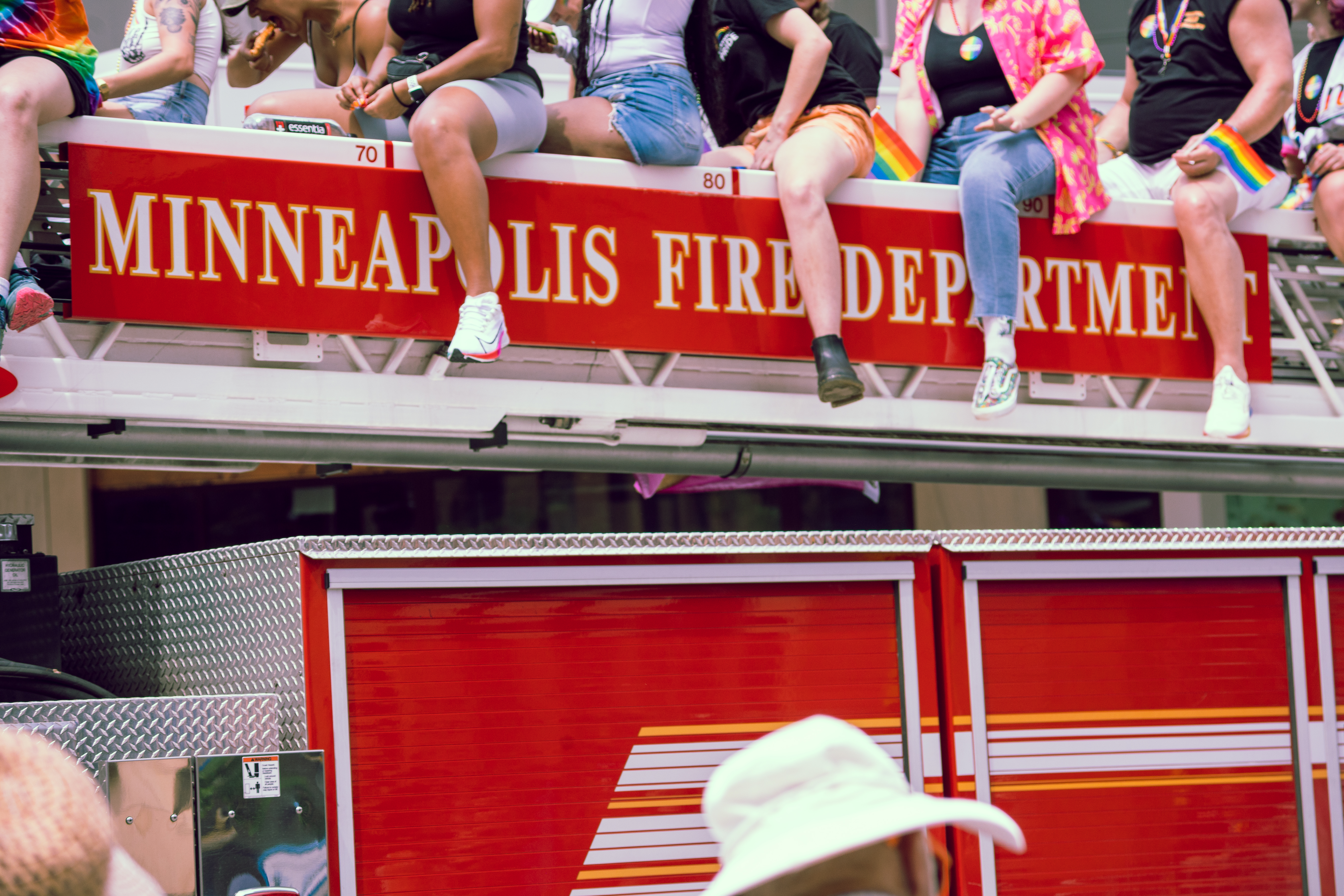 People sit on top of a Minneapolis Fire Department ladder truck during a Pride parade, smiling and waving rainbow flags.