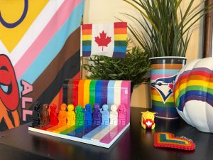 A black shelf with a collection of LGBTQ+ items: a rainbow Canadian pride flag, the Everyone is Awesome Lego set, a Pride Toronto Blue Jays cup with a green plant, a white pumpkin with a rainbow, a rainbow heart keychain, and a yellow Canadian Wapuu holding a red ball with a white maple leaf. 