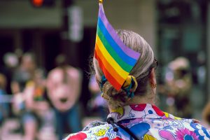 A person with light hair pulled into a bun wears a rainbow pride flag tucked into a colorful hair scarf. They are dressed in a vibrant, cartoon-printed shirt while watching a Pride parade, with a blurred crowd in the background. 
