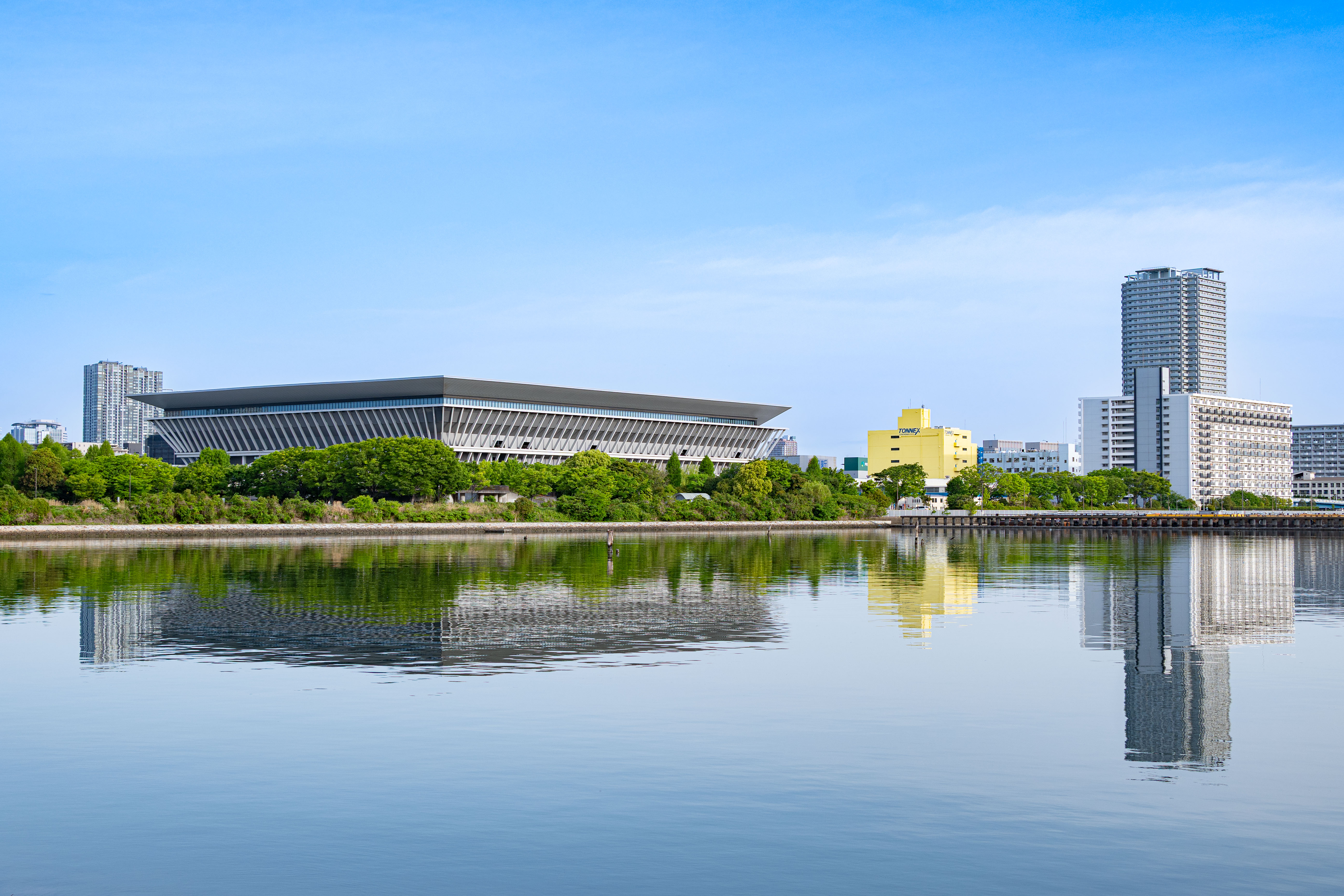 Under a clear blue sky, the Tokyo Aquatics Center and nearby apartment complexes, nestled in greenery, are beautifully reflected on the calm canal waters.