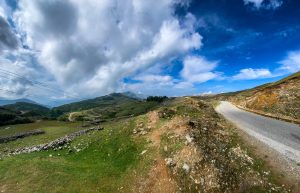 A scenic view of a winding road cutting through green hills and rocky terrain under a partly cloudy sky. 