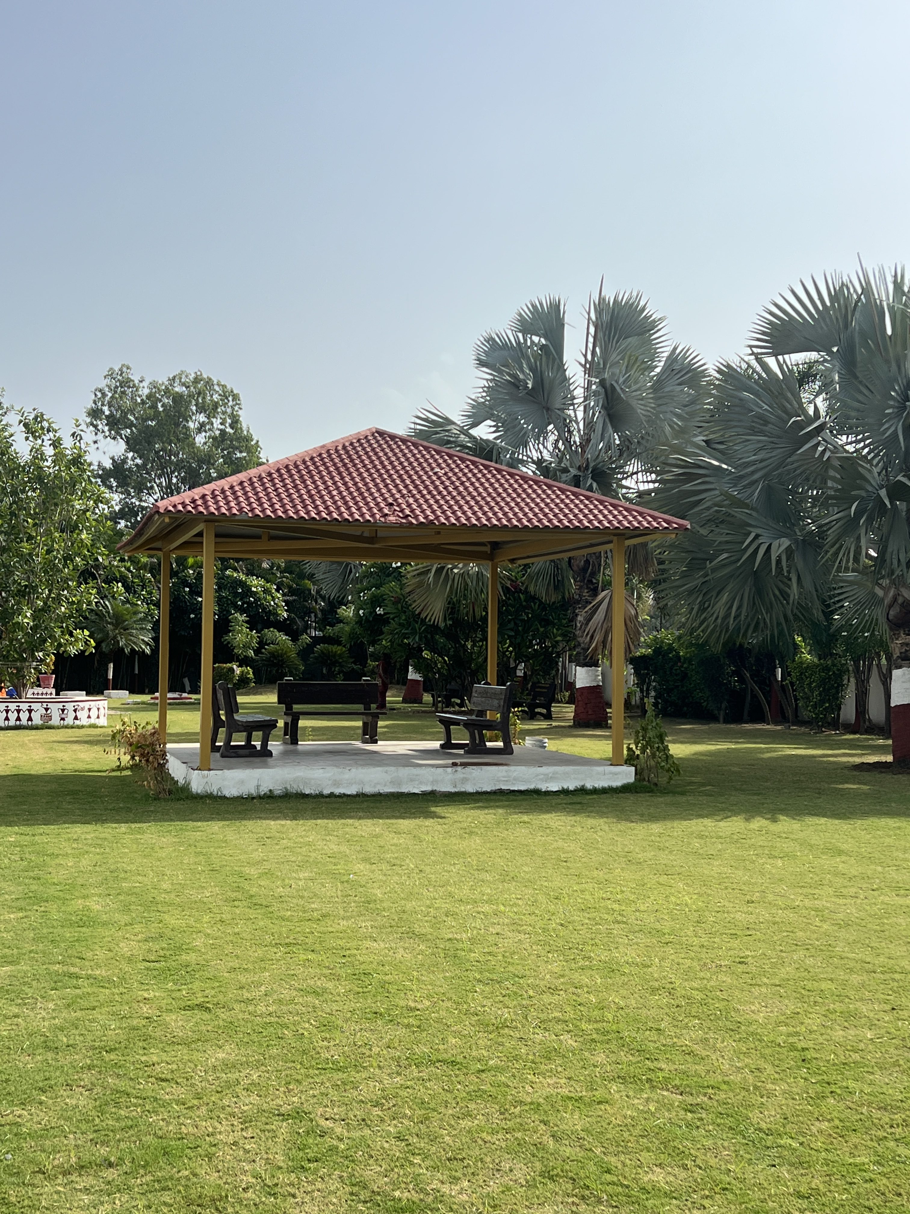 The photo shows a peaceful garden with a red-roofed gazebo in the centre. The gazebo stands on a raised white platform and contains benches and a table. It is surrounded by green grass, palm trees, and other lush greenery under a clear, sunny sky.