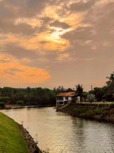 Golden hour over a riverside house in Kundapura, Karnataka, India — the warm sunset sky reflects on calm waters, painting a serene landscape embraced by lush greenery.
