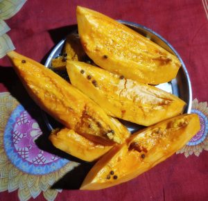 A silver plate filled with several cut pieces of ripe papaya, showcasing their bright orange flesh and black seeds. The background features a red tablecloth with decorative patterns.
