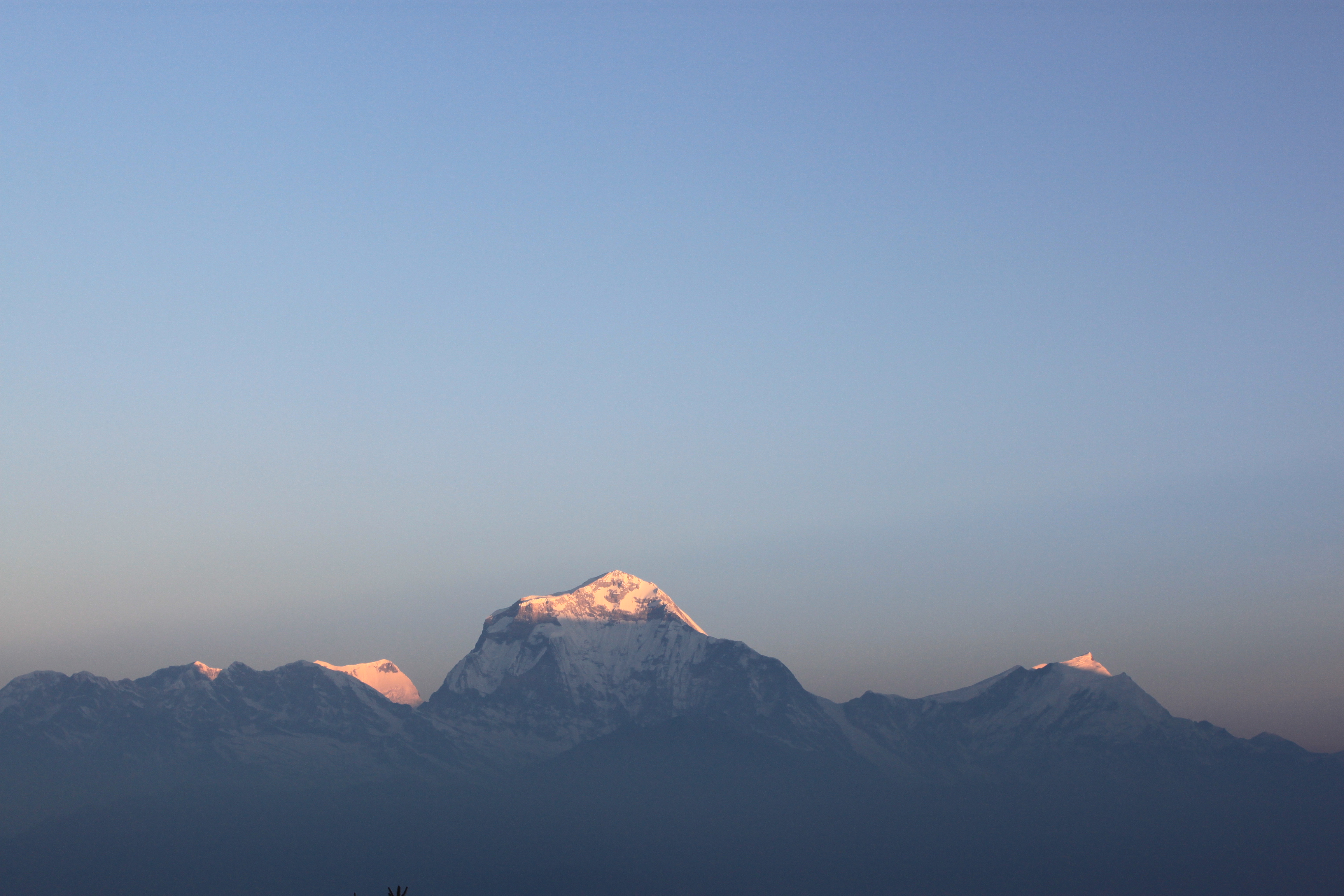 A panoramic view of rugged, snow-capped mountains under a clear blue sky during sunrise.