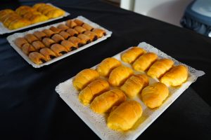 Three trays of freshly baked pastries are displayed on a black tablecloth. The front tray features golden, soft rolls arranged neatly, while the middle tray holds a variety of smaller, brown pastries,  filled with chocolate.
