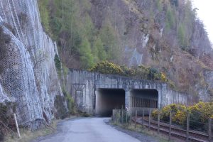 A winding road leads up to a concrete tunnel entrance carved into a hillside, flanked by a steep rock face covered with protective wire mesh. A railway track curves to the right, near the tunnel, while sturdy wooden posts demarcate the roadside. 
