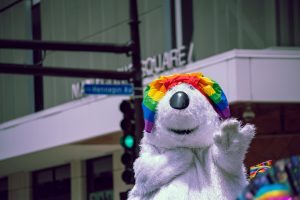 A large white polar bear waves during a Pride parade. The bear wears a rainbow-colored bandana. 
