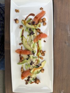 An elegant plated dish featuring slices of smoked salmon, avocado, crunchy caramelised nuts, and microgreens, artistically drizzled with balsamic and herb-infused oils on a white rectangular plate.
