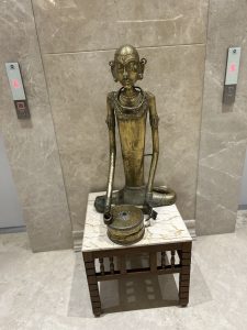 A bronze statue of a seated figure with an elongated neck and facial features, adorned with jewelry, is displayed on a marble-topped wooden table.