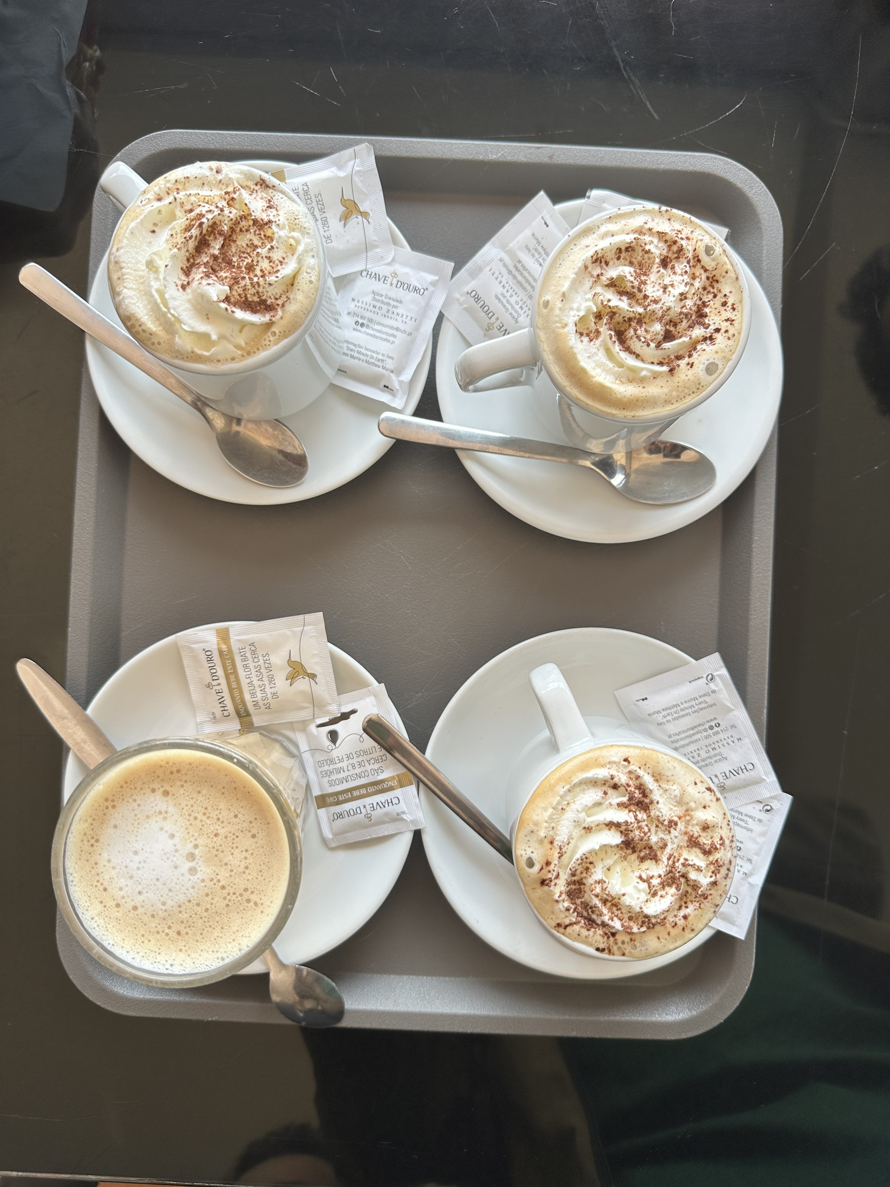 A tray of creamy, decadent coffees—three topped with swirls of whipped cream and chocolate dust, and one with a smooth frothy layer—promises a perfect pause in a Lisbon café.