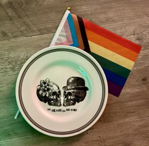 Progress pride flag on a wood table that has a plate sitting on top which has a design of a skull with a flower facing a skull with bowler hat above the words til death do us part.