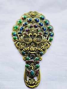 An ornate antique metal mirror and comb set adorned with peacock motifs and colorful gem-like stones.