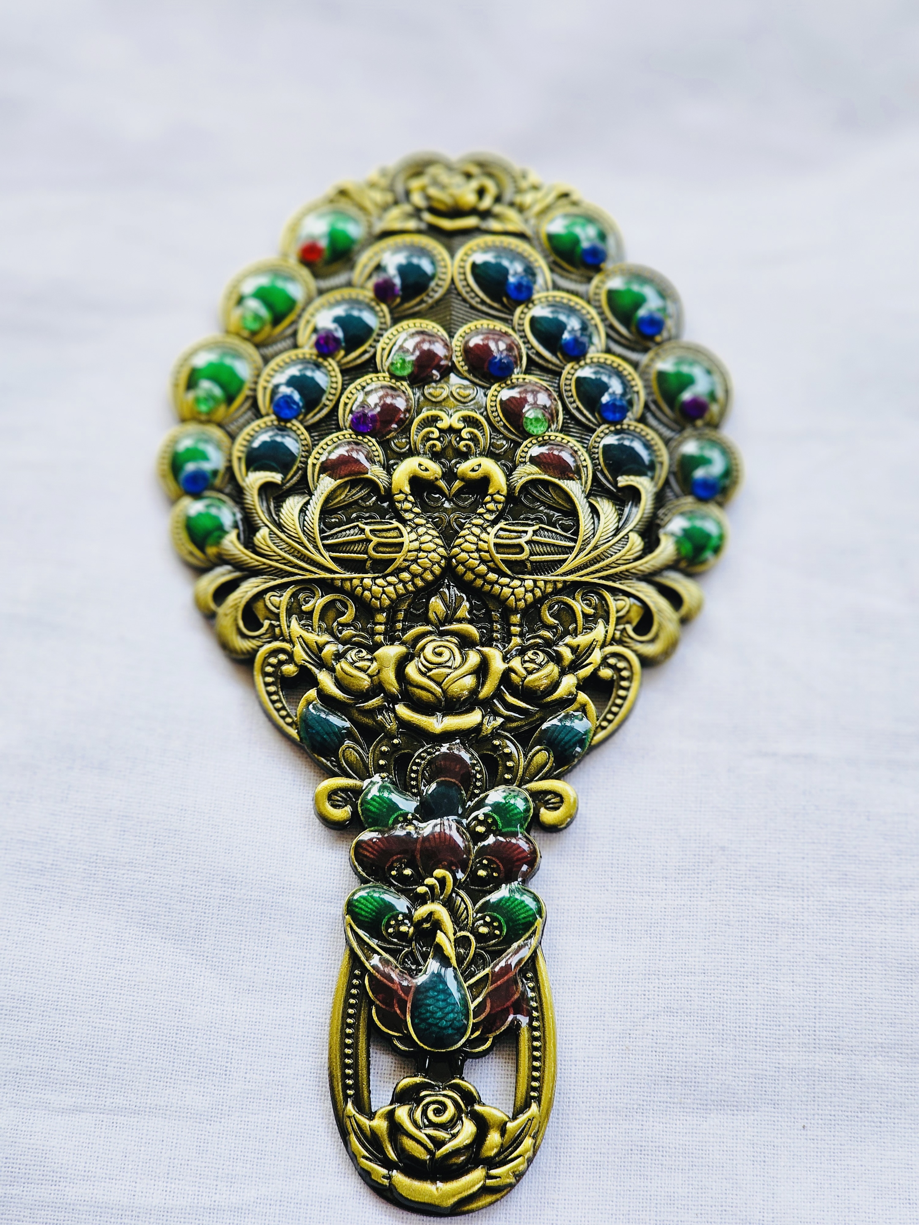 An ornate antique metal mirror and comb set adorned with peacock motifs and colorful gem-like stones.