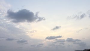 A serene sky at dusk, filled with soft, fluffy clouds in various shades of gray and white, set against a pale blue background. The light is gentle and diffused, creating a calm and tranquil atmosphere.