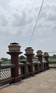 A view of a riverside walkway with decorative stone pillars. The pillars are connected by a railing with a crisscross metal design.