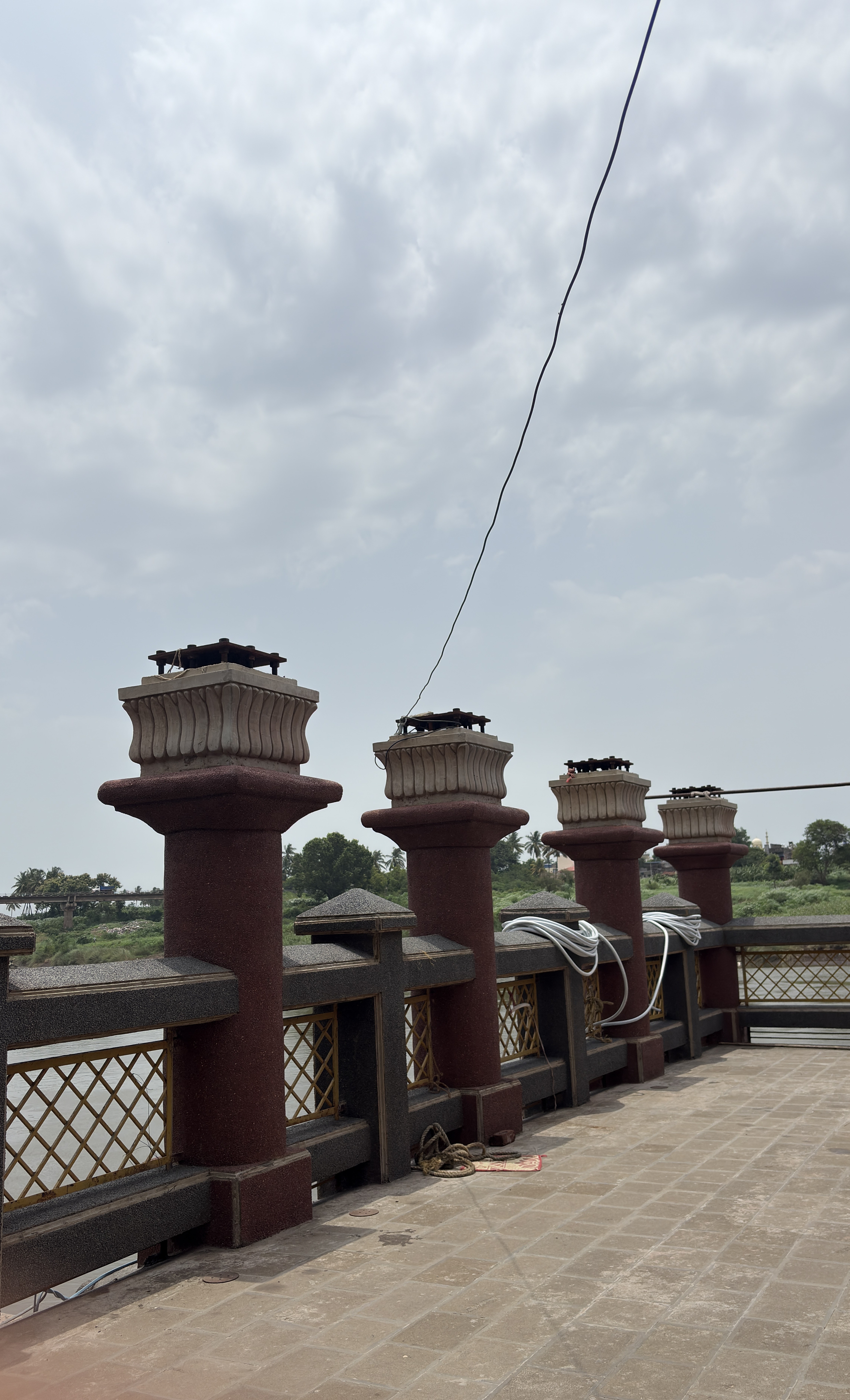 A view of a riverside walkway with decorative stone pillars. The pillars are connected by a railing with a crisscross metal design.