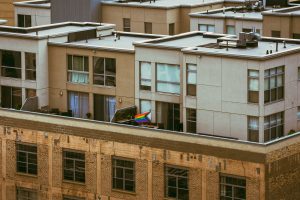 Looking down on a rooftop apartment patio displays a Progress Pride flag waving in the wind. The building has large windows and beige walls, with an older brick structure visible in the foreground. 