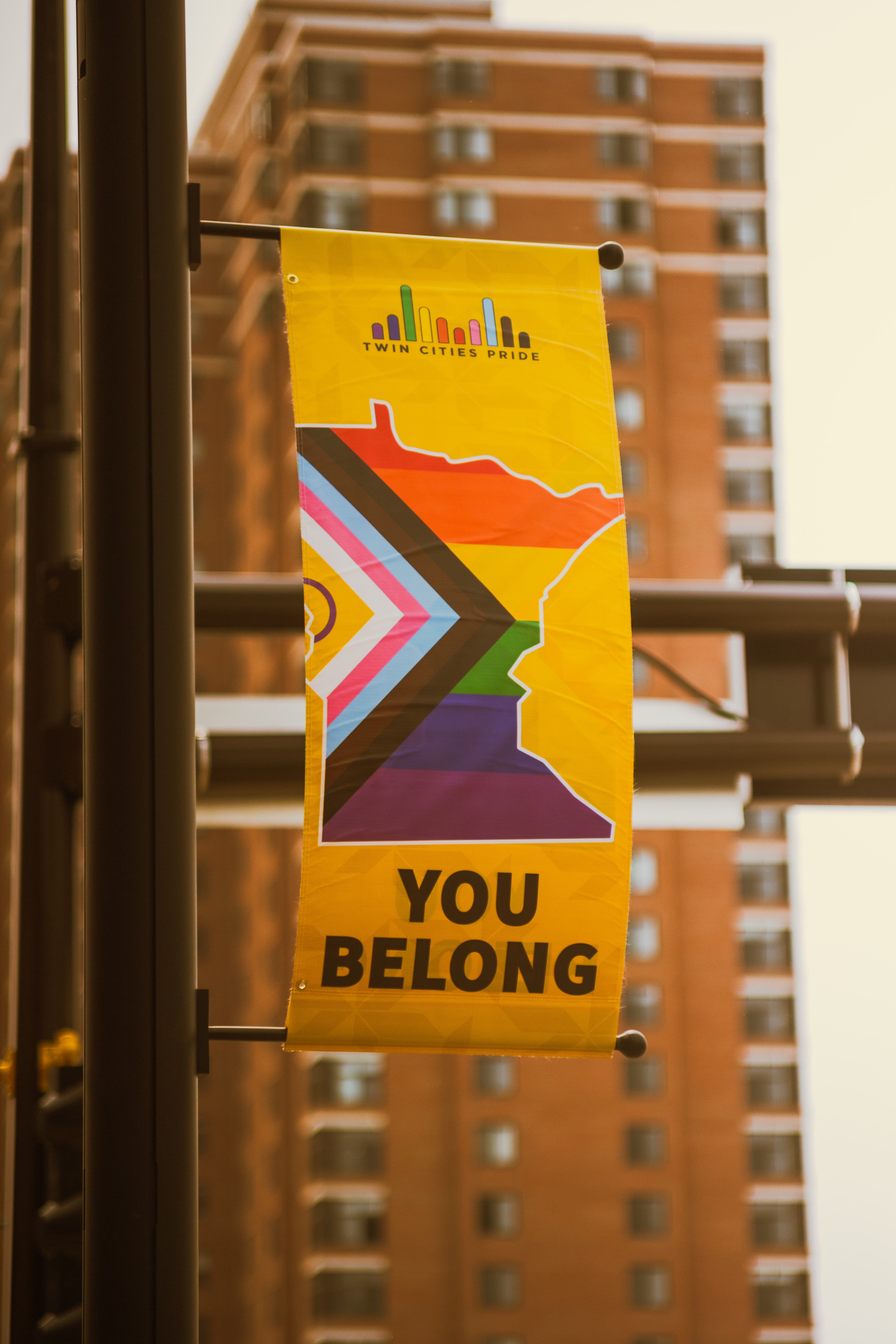 A yellow banner hangs from a light poll with "Twin Cities Pride" and a rainbow bridge logo at the top, the outline of the state of Minnesota filled in with the progress pride flag and "You Belong" at the bottom. In the background is a tall brick building.