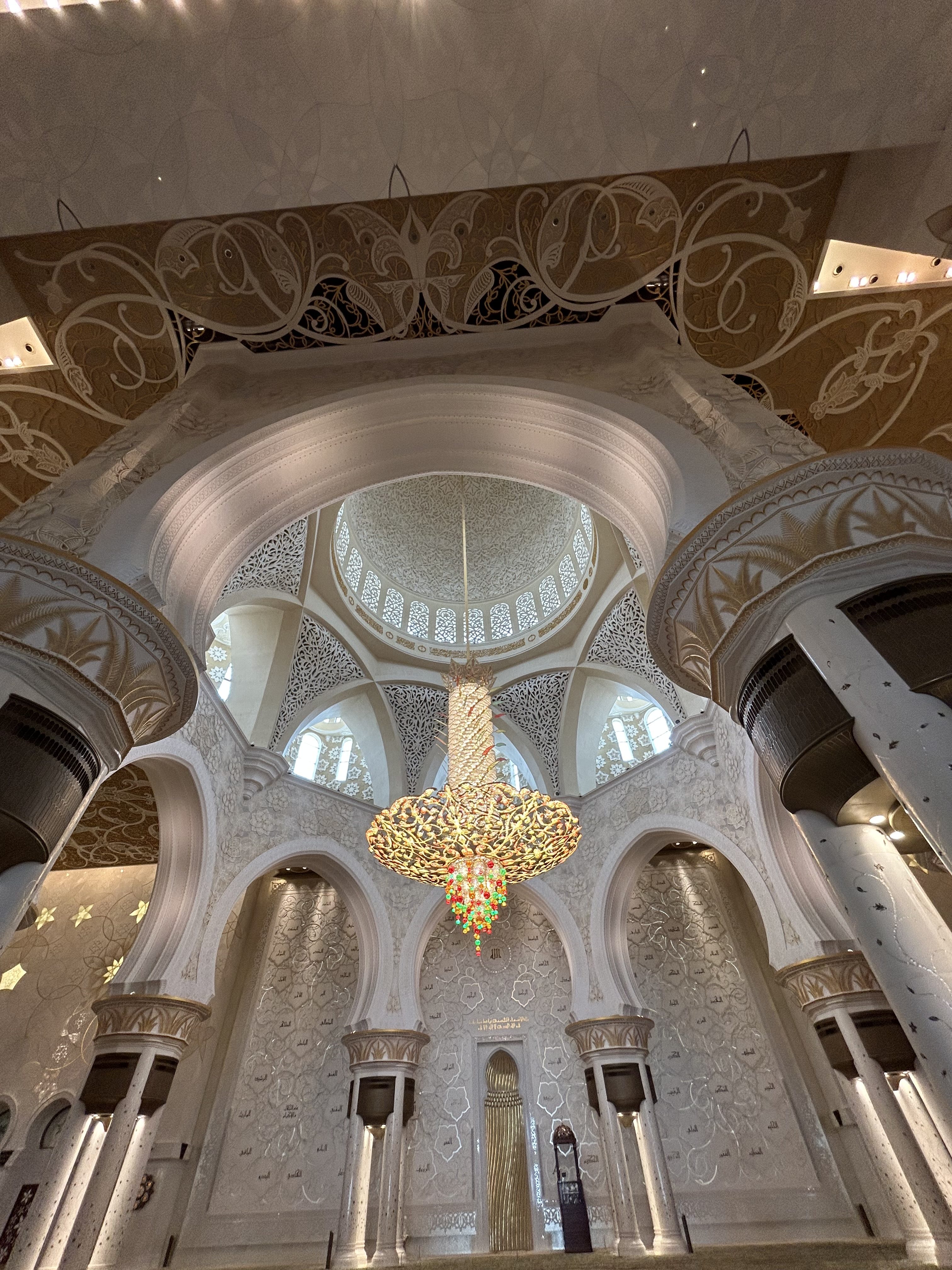 Interior of an ornate mosque with white marble columns, intricate Islamic patterns, and a large golden chandelier hanging from the domed ceiling.