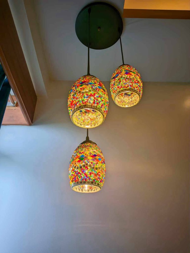 Three colorful, mosaic-style pendant lights hang from a dark ceiling mount against a plain wall. Each light fixture features intricate patterns made from small, vibrant glass pieces, emitting a warm glow.