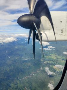 A view from an airplane window featuring a large black propeller with distinct blades, set against a backdrop of blue sky and scattered white clouds.