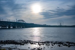 In early morning light, the sun reflects on the Arakawa River, with the JR Keiyo Line bridge and a distant Ferris wheel at the river's mouth.