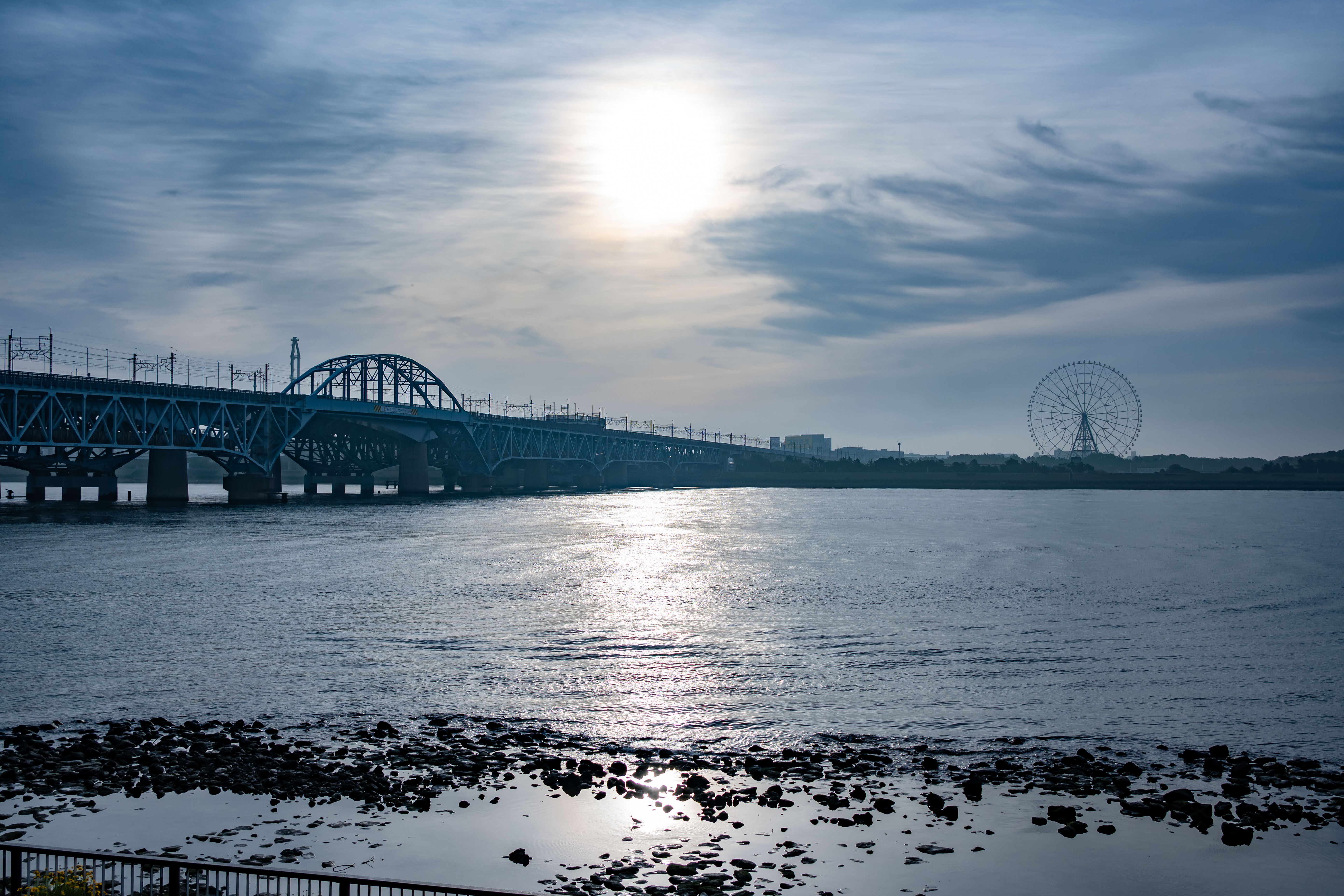 In early morning light, the sun reflects on the Arakawa River, with the JR Keiyo Line bridge and a distant Ferris wheel at the river's mouth.