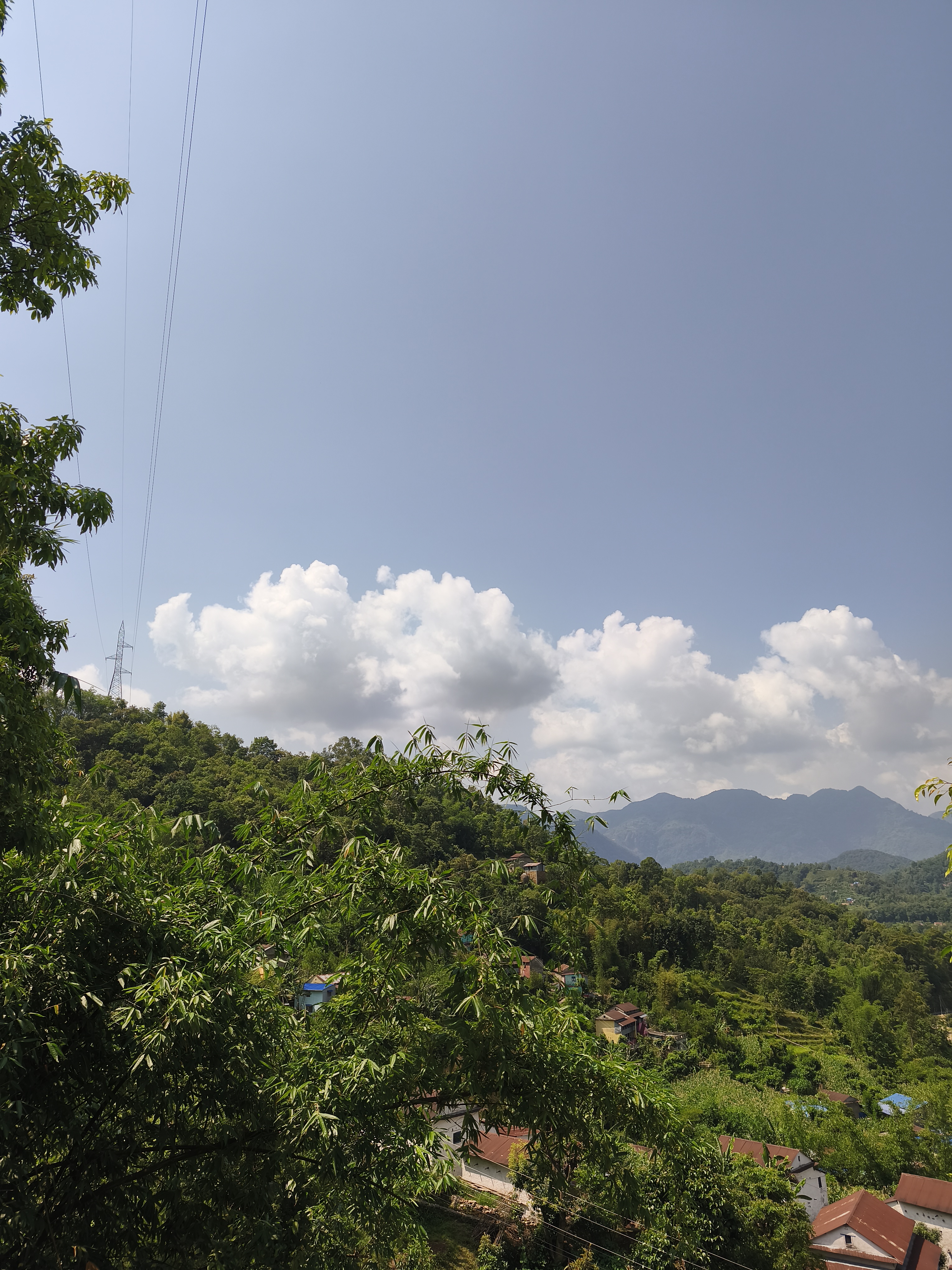 A scenic view of a lush green landscape, featuring rolling hills and mountains in the background under a clear blue sky with scattered white clouds.