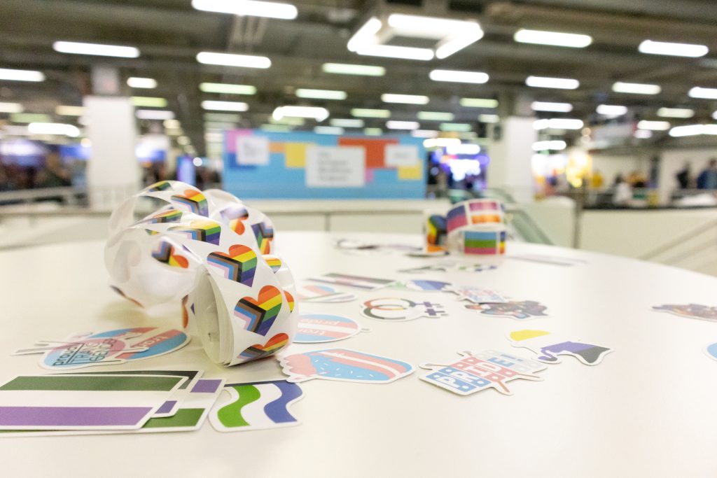 Close-up of a white table covered in various LGBTQIA+ pride stickers, including rainbow hearts, progress flags, and symbols for gender and sexual diversity. In the background, a colorful community banner is slightly out of focus, hinting at an event or conference setting with bright lights and people mingling.