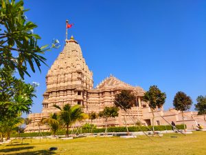 Khodaldham Temple in Kagvad, Rajkot, Gujarat, features light stone, tall carved towers, and a red flag atop the main shikhar, set amid green lawns, palm trees, and a bright blue sky.