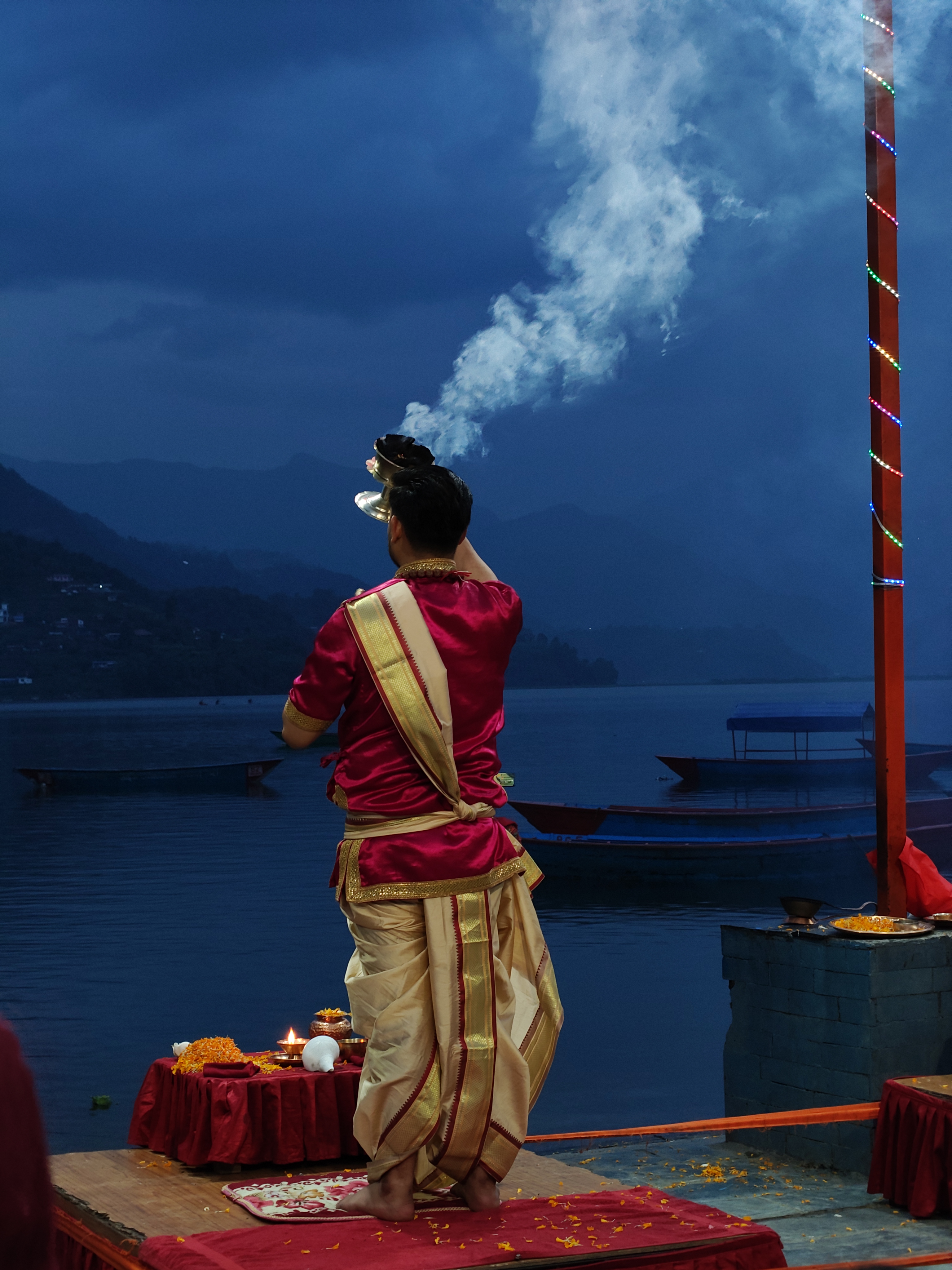 A person in traditional attire is standing by a lake, facing away from the camera.