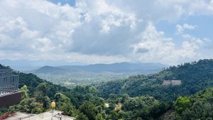 A panoramic view of lush green hills and valleys under a partly cloudy sky.
