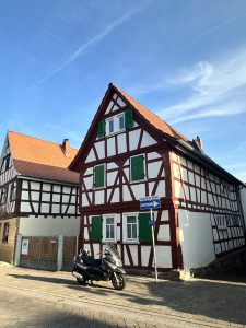 A charming half-timbered house with green shutters and a red-tiled roof sits on a cobbled corner under a clear blue sky, with a parked scooter adding a modern touch to the historic street scene.