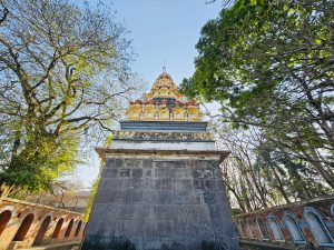 Backyard of Kartik Swami Temple at Parvati Hill, Pune, with a colorful tiered tower, stone base, trees, and arched brick wall