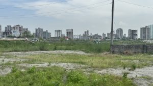 A grassy field with patches of sand and unfinished buildings in the background.