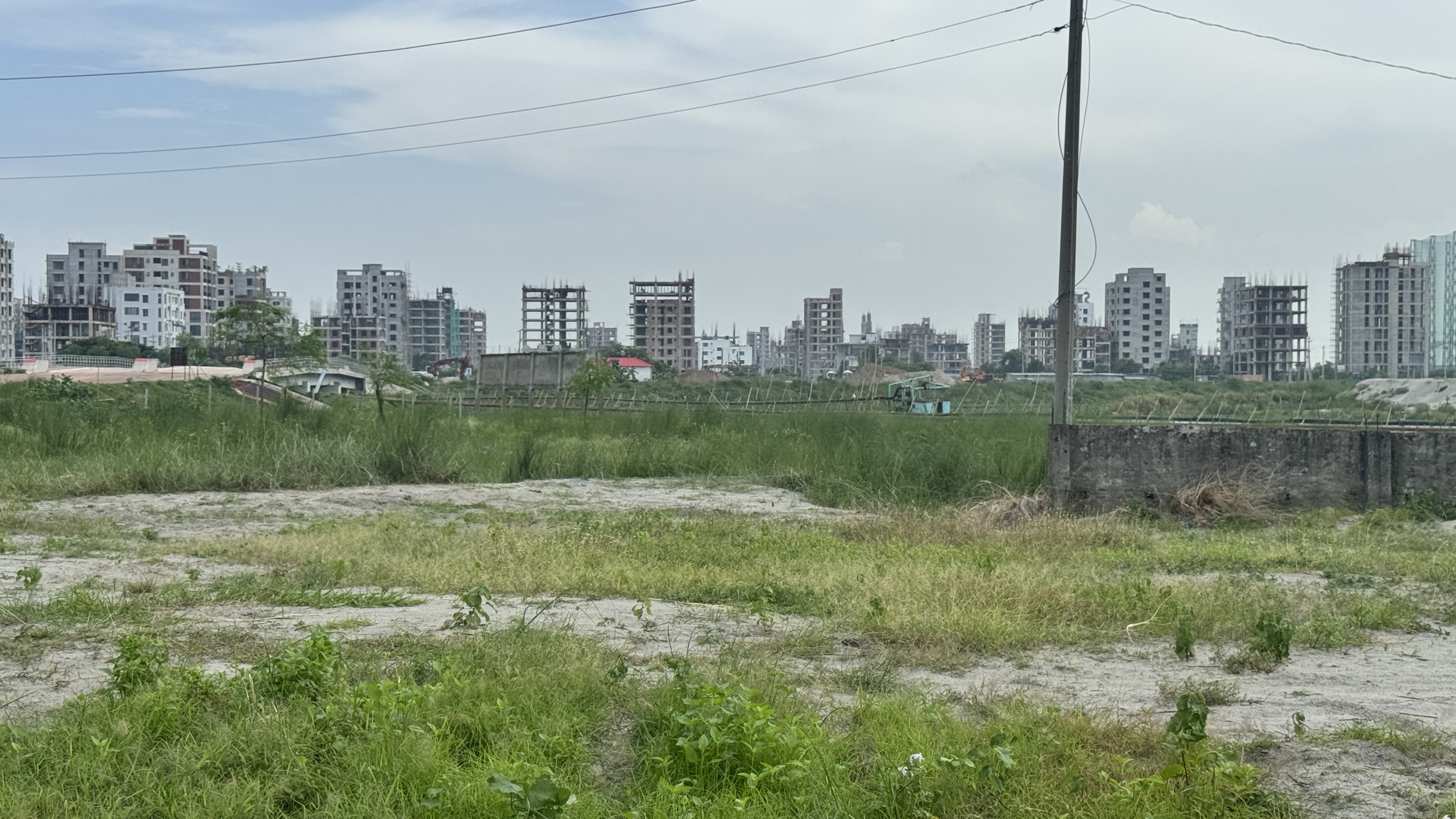 A grassy field with patches of sand and unfinished buildings in the background.