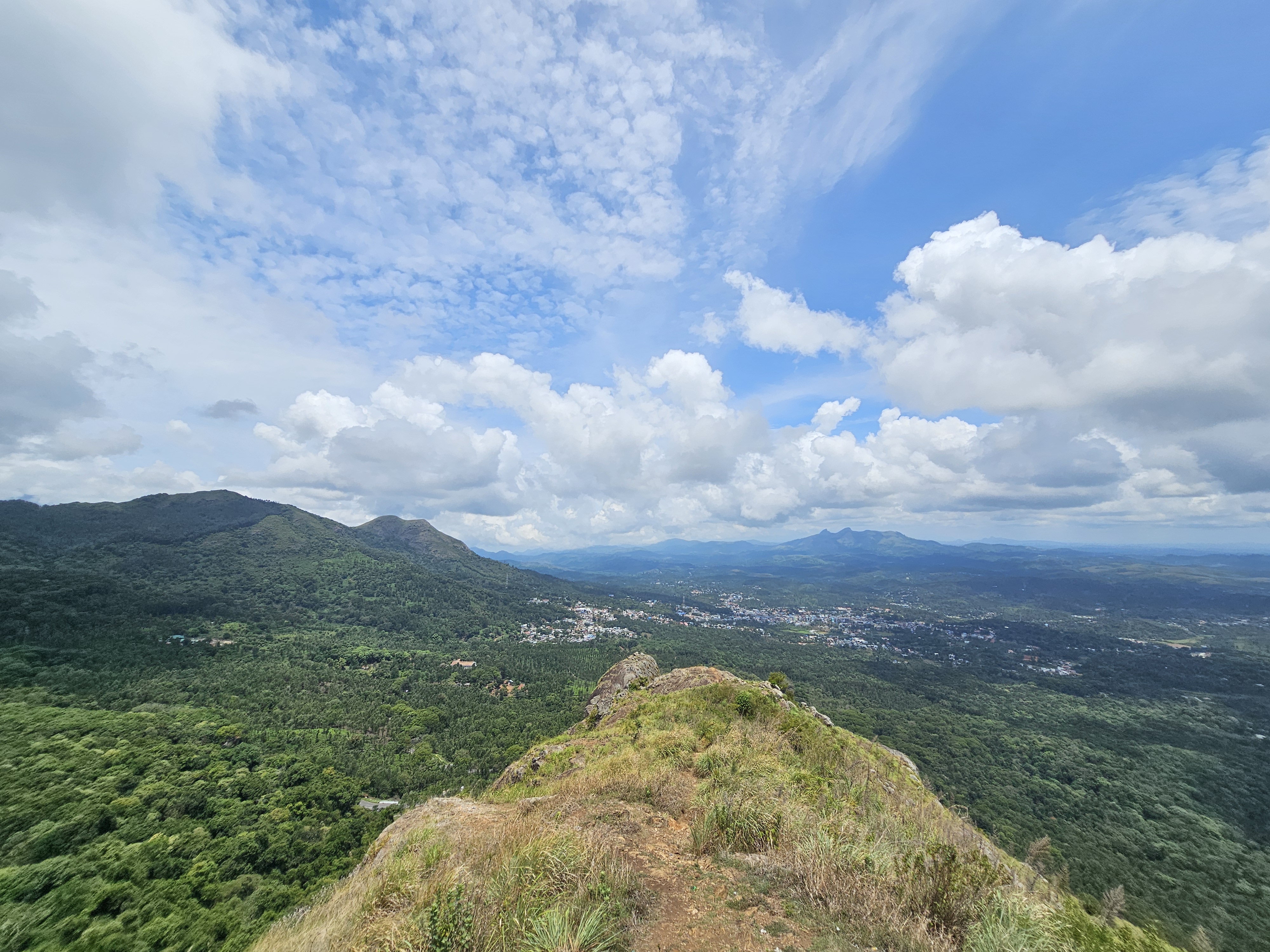 A scenic hilltop view from Needle Rock View Point, Gudalur, showcases lush Nilgiri mountains and a distant town under a partly cloudy sky.